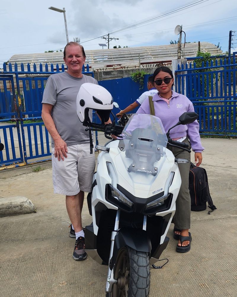 Traveller and a smiling woman with a white Honda ADV 160 by the blue airport fence