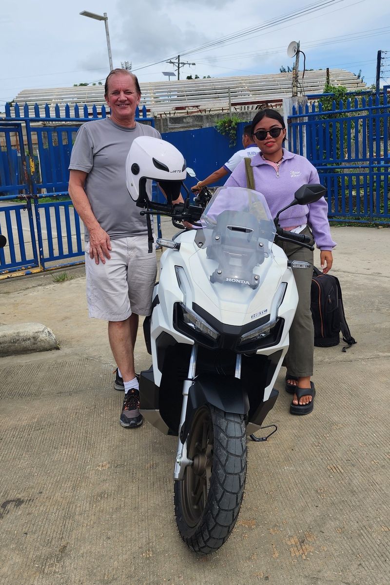 Traveller and a smiling woman with a white Honda ADV 160 by the blue airport fence