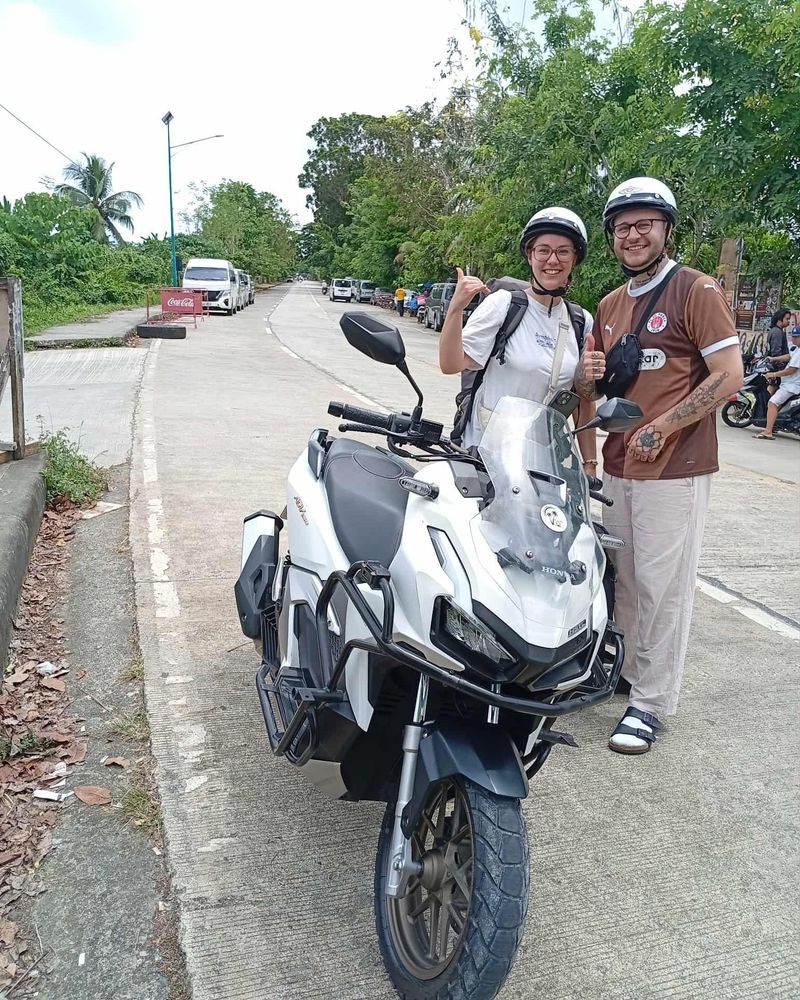 Smiling couple in helmets giving thumbs-up beside a white Honda ADV 160