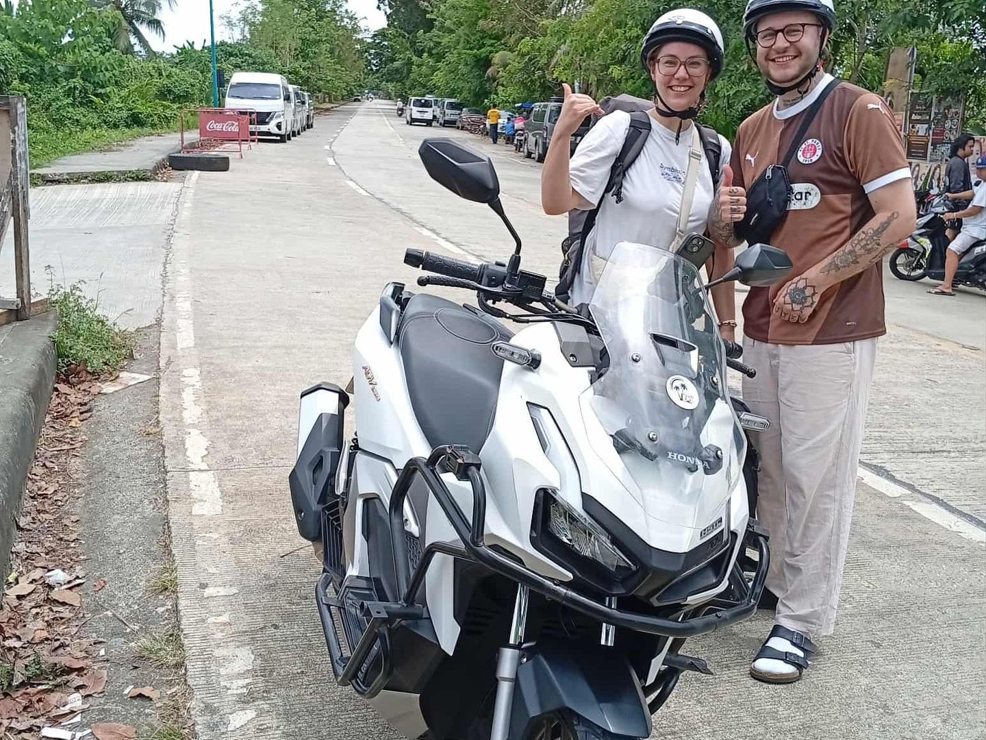 Smiling couple in helmets giving thumbs-up beside a white Honda ADV 160