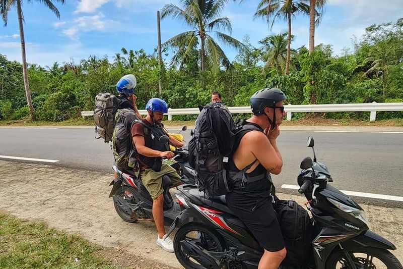 Backpackers in helmets setting off on Honda BeAT scooters along a palm-lined road