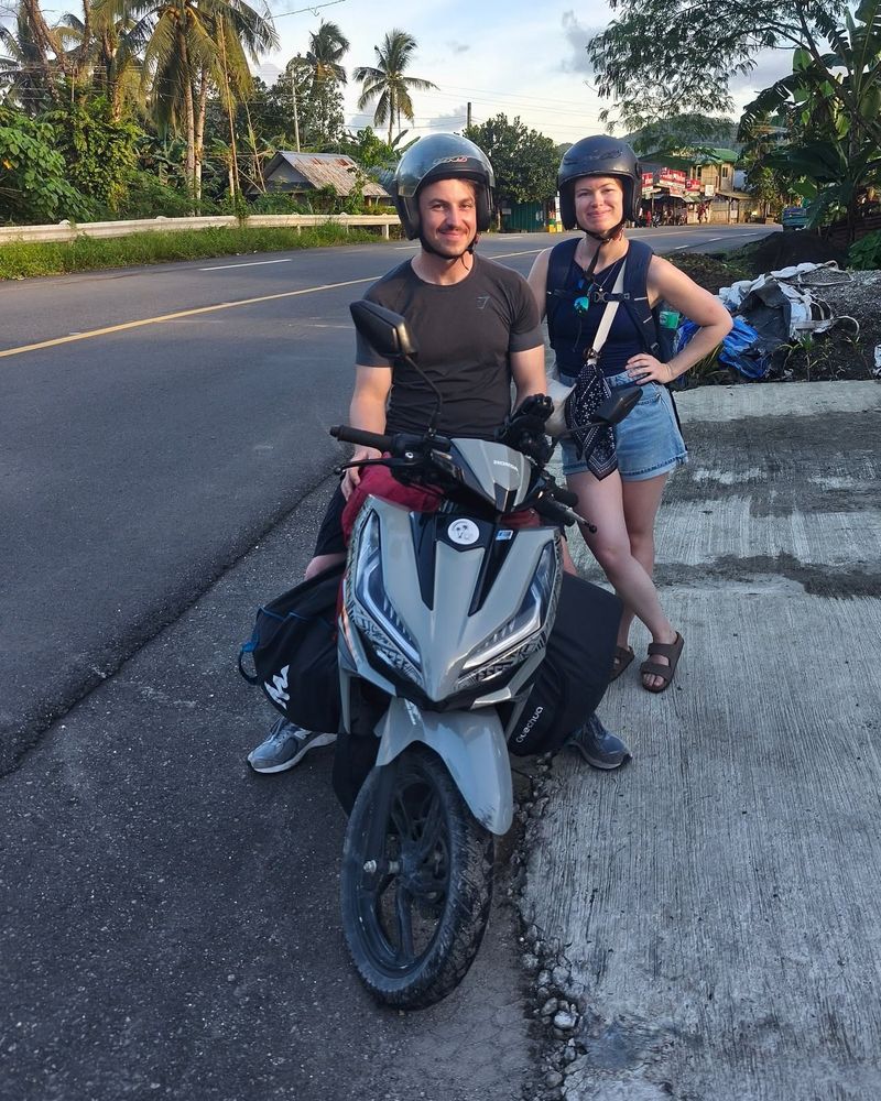Smiling couple in helmets with a grey Honda Click on a highway in late-afternoon light
