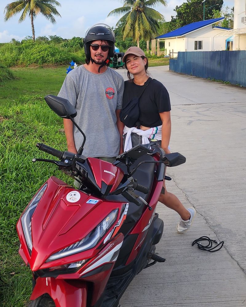 Couple standing behind a red Honda Click scooter on a palm-lined road