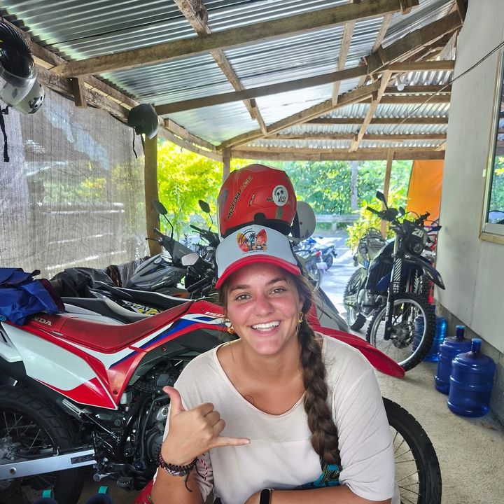 Smiling guest in a Siargao Rose cap giving a shaka at the shop desk, with dirt bikes behind
