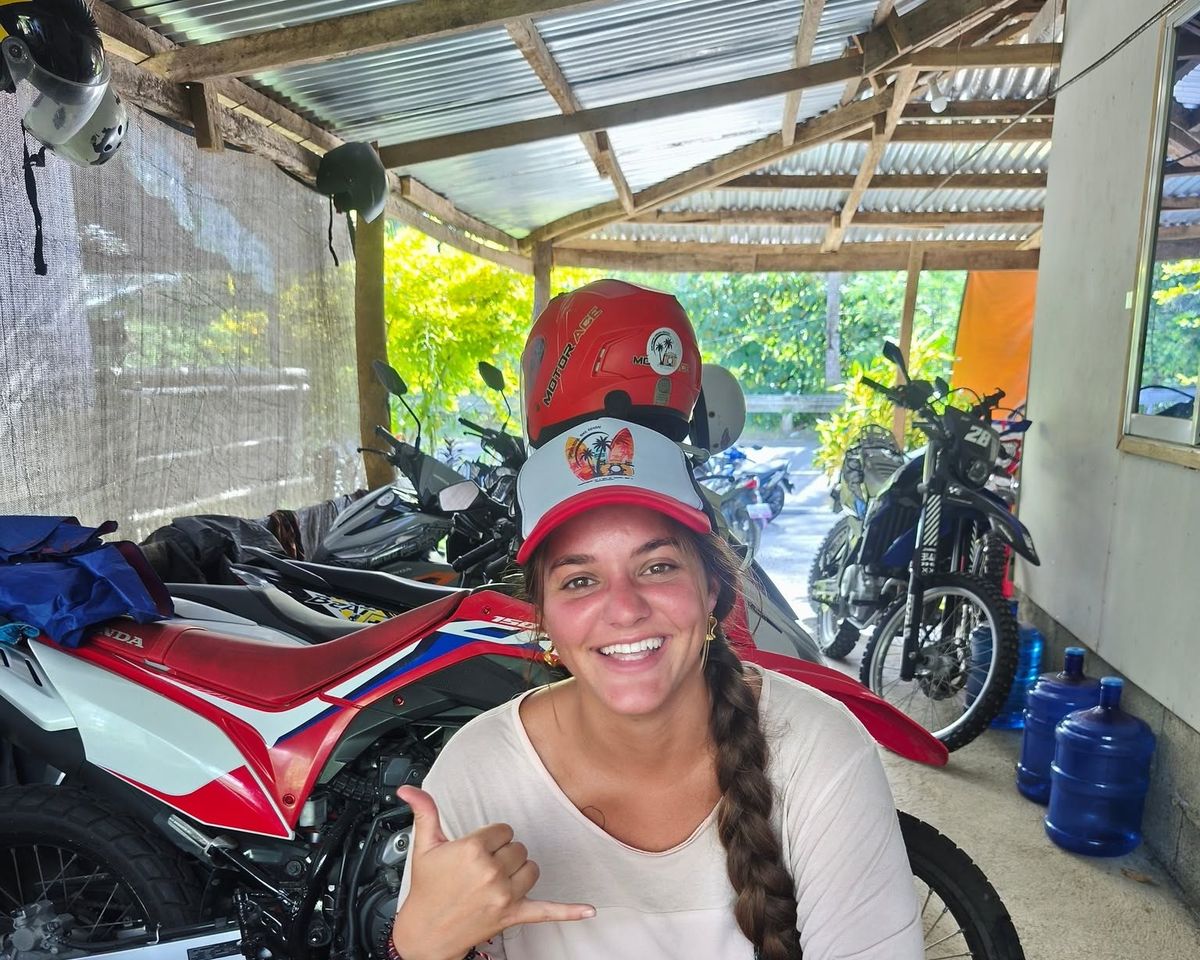 Smiling guest in a Siargao Rose cap giving a shaka at the shop desk, with dirt bikes behind