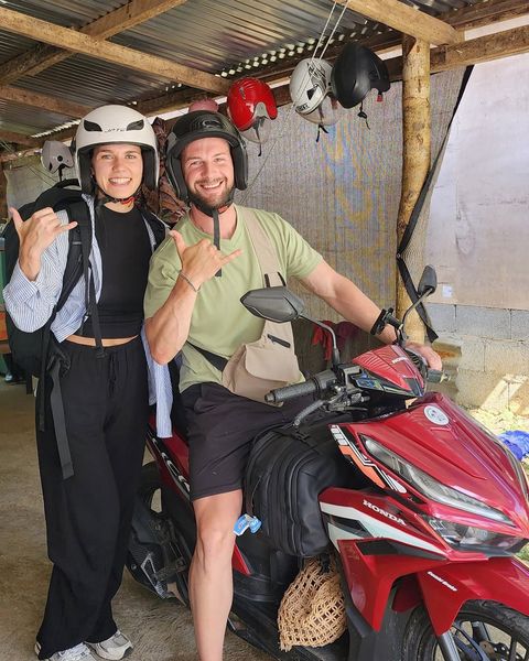 Two smiling guests in helmets giving shakas on a red Honda scooter under the rental shop shed