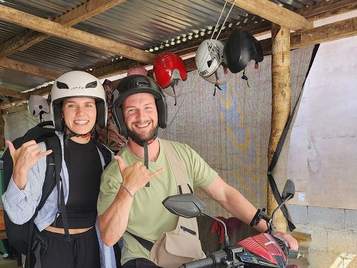 Two smiling guests in helmets giving shakas on a red Honda scooter under the rental shop shed
