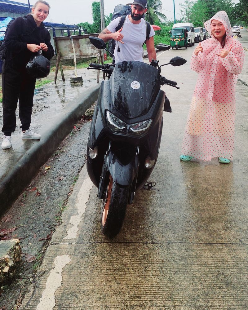 Travellers and a host in a polka-dot raincoat beside a black Yamaha NMAX on a rainy day