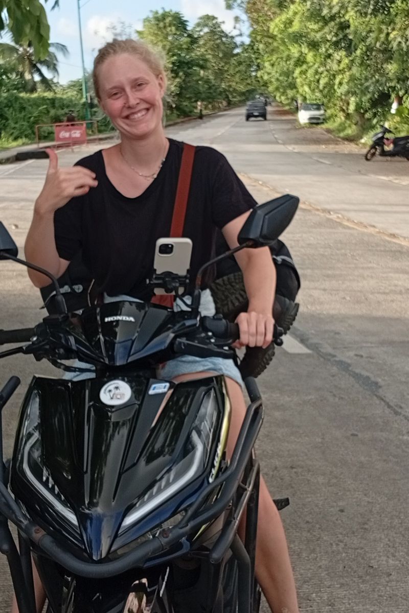 Smiling rider giving a shaka on a black Honda Click with crash bars on a tree-lined road