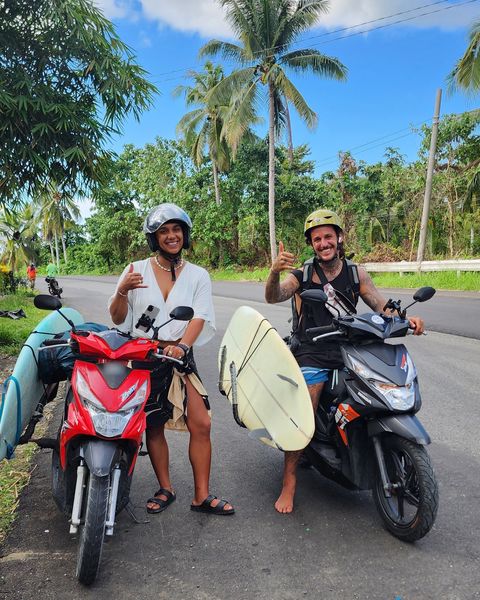 Two smiling riders in helmets on rental scooters with surfboards in side racks on a palm-lined road