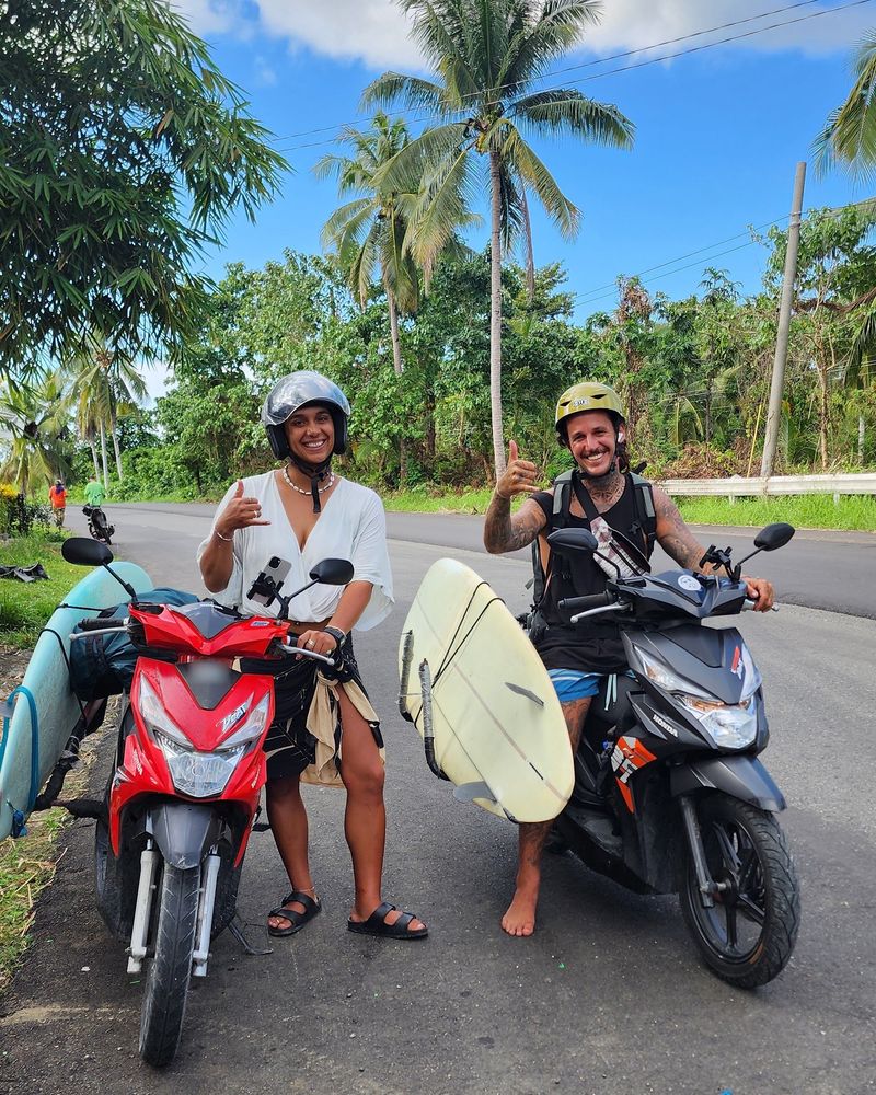 Two smiling riders in helmets on rental scooters with surfboards in side racks on a palm-lined road