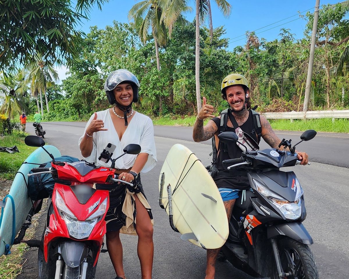 Two smiling riders in helmets on rental scooters with surfboards in side racks on a palm-lined road