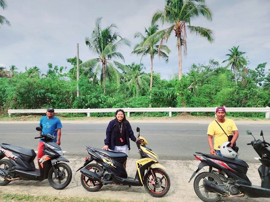 Three members of the Siargao Rose team standing behind rental scooters on a palm-lined highway