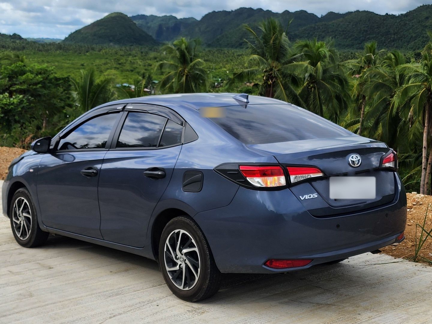 Blue-grey Toyota Vios sedan parked on a hilltop above jungle-covered hills