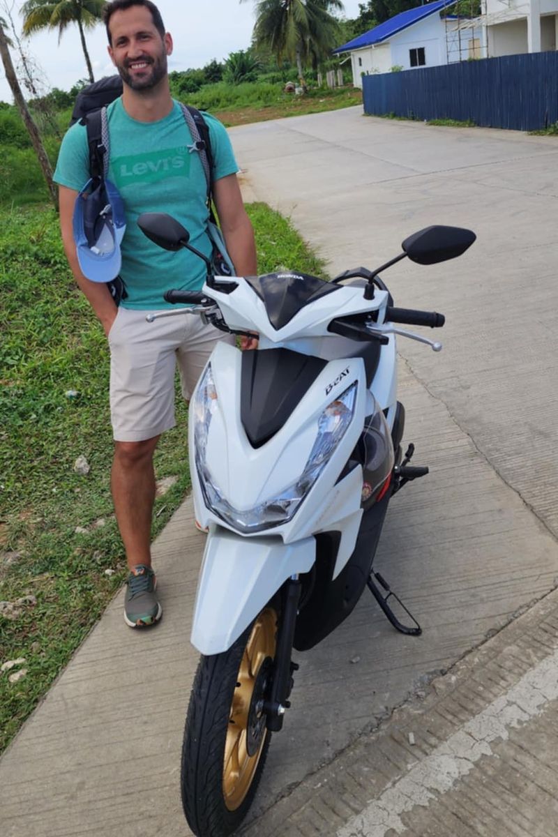 Traveller standing beside a white Honda BeAT with gold wheels on a palm-lined street