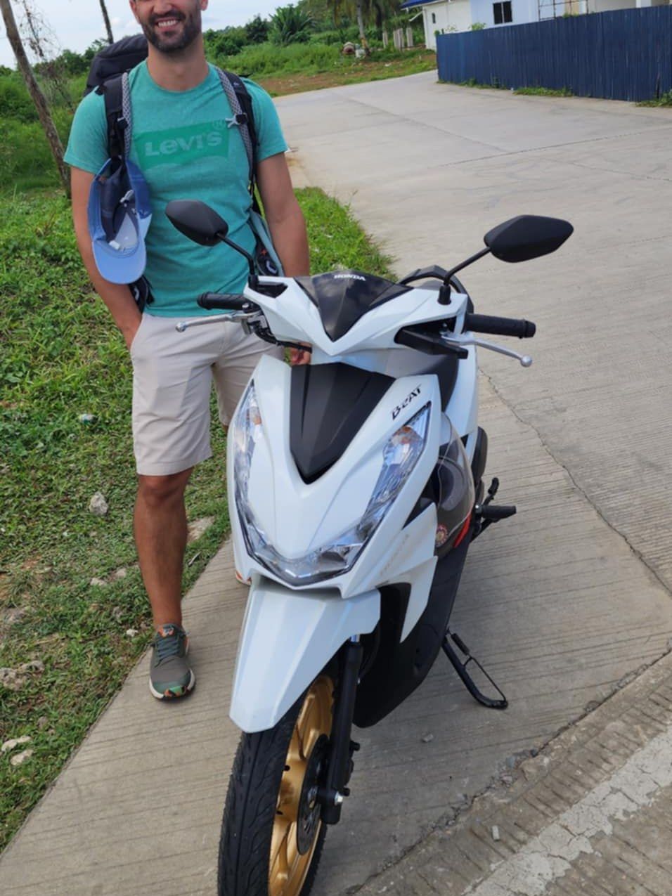 Traveller standing beside a white Honda BeAT with gold wheels on a palm-lined street