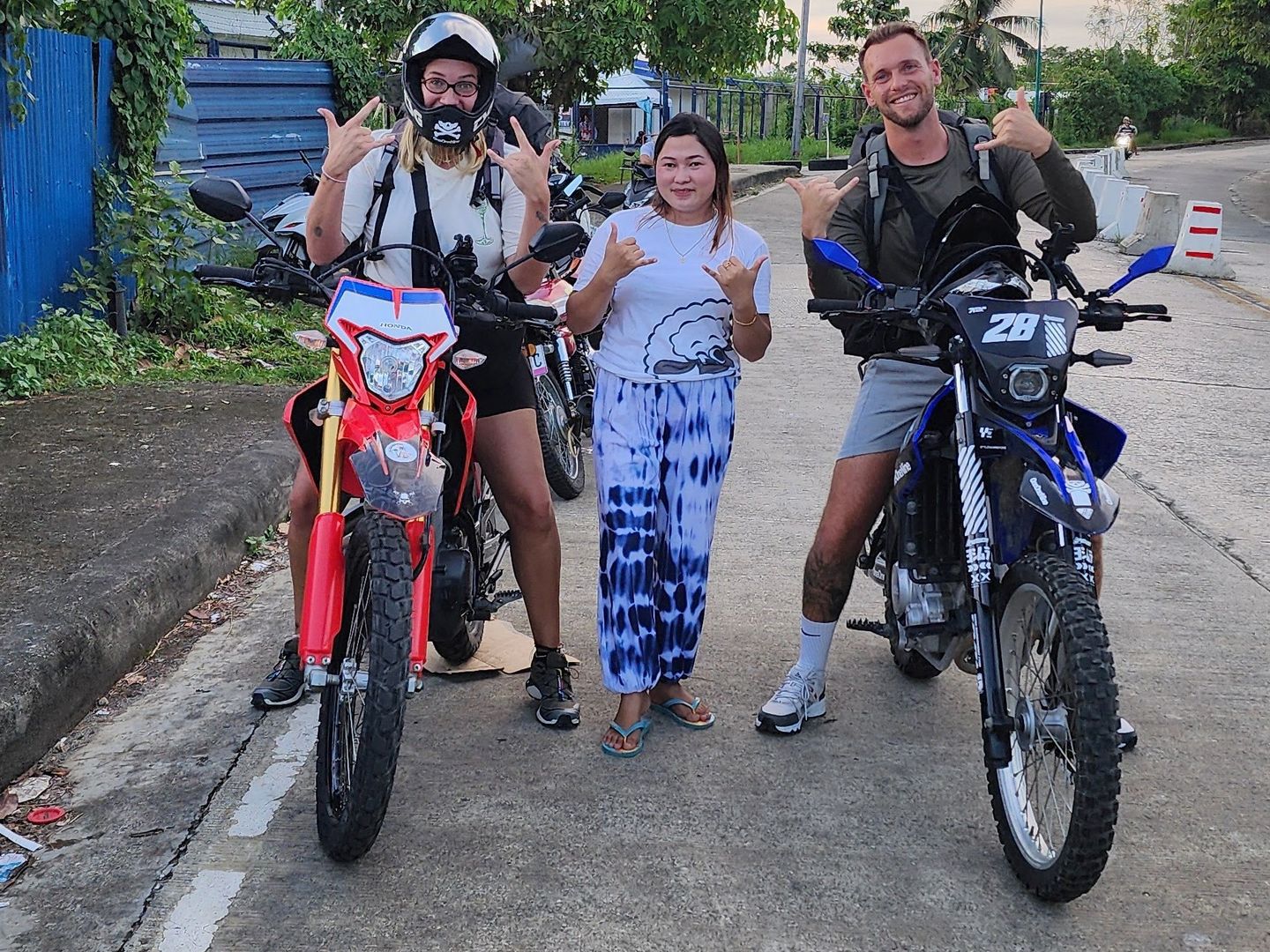 A traveller in a full-face helmet with a red Honda CRF150L and a bareheaded traveller with a blue Yamaha WR155R, a smiling woman standing between them